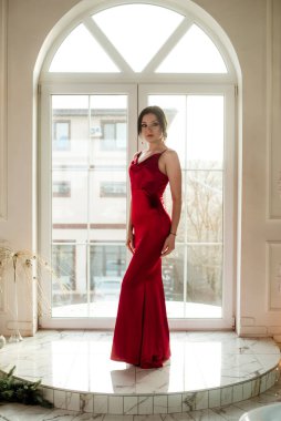 Siyah saçlı, gece makyajlı, uzun kırmızı bir gece elbisesi ve büyük vitraylı pencereli çekici bir genç kız. Güzellik ve moda 