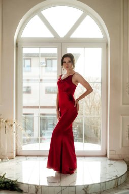 Siyah saçlı, gece makyajlı, uzun kırmızı bir gece elbisesi ve büyük vitraylı pencereli çekici bir genç kız. Güzellik ve moda 