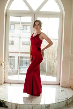 Siyah saçlı, gece makyajlı, uzun kırmızı bir gece elbisesi ve büyük vitraylı pencereli çekici bir genç kız. Güzellik ve moda 