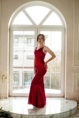 Siyah saçlı, gece makyajlı, uzun kırmızı bir gece elbisesi ve büyük vitraylı pencereli çekici bir genç kız. Güzellik ve moda 