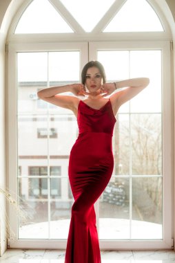 Siyah saçlı, gece makyajlı, uzun kırmızı bir gece elbisesi ve büyük vitraylı pencereli çekici bir genç kız. Güzellik ve moda 