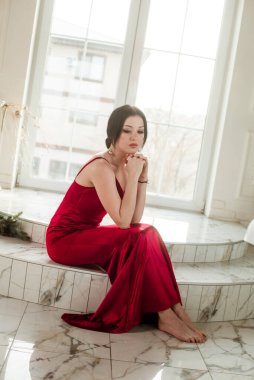 Siyah saçlı, gece makyajlı, uzun kırmızı bir gece elbisesi ve büyük vitraylı pencereli çekici bir genç kız. Güzellik ve moda 