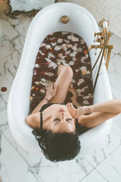 バラの花びらでいっぱいの浴槽に黒いビキニで暗い髪を持つ魅力的な若い女の子 レジャー — ストック写真