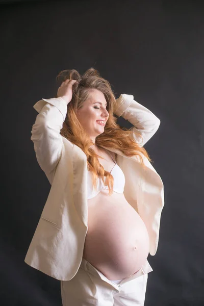 黒の背景に白いズボンのスーツに長い赤い髪の若いかわいい妊娠中の女性 幸せな母親 母の日 — ストック写真