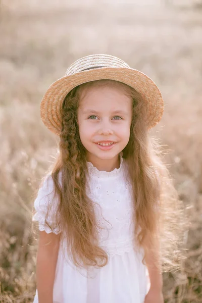 一个可爱的小女孩 留着一头长长的金发卷发 穿着白色的夏装 夕阳西下 在乡间的田野里戴着一顶稻草帽 自然与生态 — 图库照片