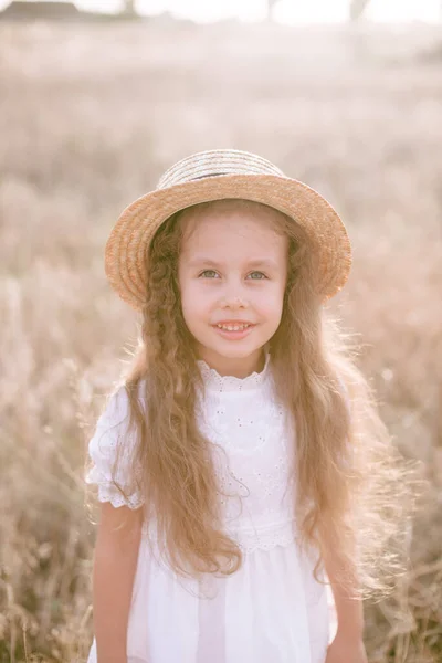 一个可爱的小女孩 留着一头长长的金发卷发 穿着白色的夏装 夕阳西下 在乡间的田野里戴着一顶稻草帽 自然与生态 — 图库照片