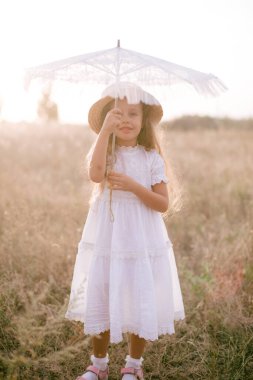 Yazın gün batımında kırsalda bir tarlada beyaz bir yaz elbisesi içinde uzun sarı kıvırcık saçlı tatlı küçük bir kız. Doğa ve Çevre Yaşamı
