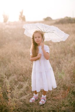 Yazın gün batımında kırsalda bir tarlada beyaz bir yaz elbisesi içinde uzun sarı kıvırcık saçlı tatlı küçük bir kız. Doğa ve Çevre Yaşamı