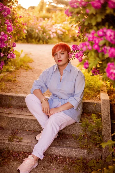 夏至时分 一位穿着蓝色时髦衬衫 穿着白色长裤 头戴红头发的年轻女子来到了玫瑰园 — 图库照片