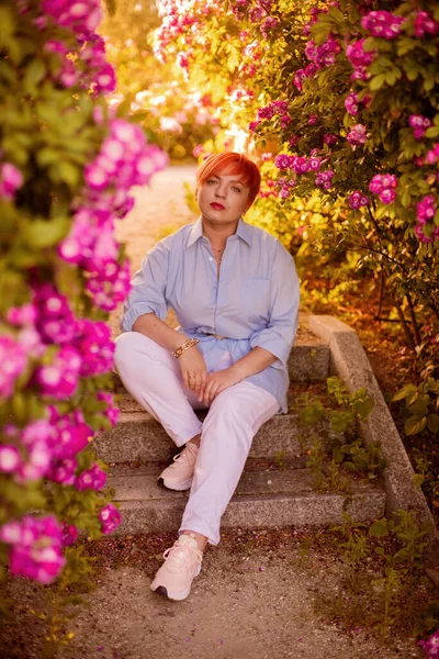 青い流行のシャツを着た赤い髪の若い女性と 夏の日没時に開花しているバラ園の白いズボン — ストック写真