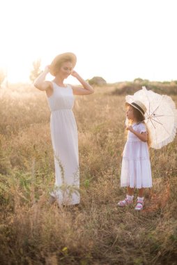 Uzun sarı kıvırcık saçlı şirin bir kız ve yaz günü güneş batarken kırsalda saman kayıkçı şapkalı beyaz elbiseli annesi. Doğa ve Çevre Yaşamı