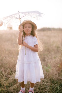 Yazın günbatımında kırsalda beyaz dantel şemsiyeli, uzun sarı kıvırcık saçlı, beyaz bir şapkası olan küçük şirin bir kız. Doğa ve Çevre Yaşamı
