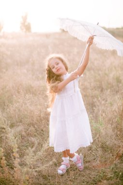 Yazın günbatımında kırsalda beyaz dantel şemsiyeli, uzun sarı kıvırcık saçlı, beyaz bir şapkası olan küçük şirin bir kız. Doğa ve Çevre Yaşamı