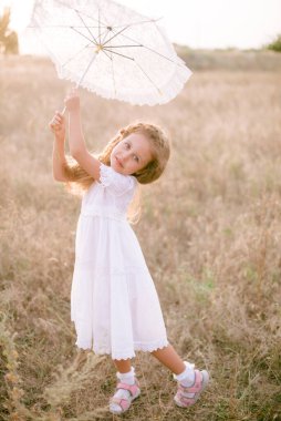 Yazın günbatımında kırsalda beyaz dantel şemsiyeli, uzun sarı kıvırcık saçlı, beyaz bir şapkası olan küçük şirin bir kız. Doğa ve Çevre Yaşamı