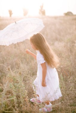 Yazın günbatımında kırsalda beyaz dantel şemsiyeli, uzun sarı kıvırcık saçlı, beyaz bir şapkası olan küçük şirin bir kız. Doğa ve Çevre Yaşamı