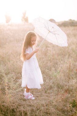 Yazın günbatımında kırsalda beyaz dantel şemsiyeli, uzun sarı kıvırcık saçlı, beyaz bir şapkası olan küçük şirin bir kız. Doğa ve Çevre Yaşamı