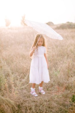 Yazın günbatımında kırsalda beyaz dantel şemsiyeli, uzun sarı kıvırcık saçlı, beyaz bir şapkası olan küçük şirin bir kız. Doğa ve Çevre Yaşamı
