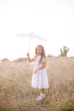 Yazın günbatımında kırsalda beyaz dantel şemsiyeli, uzun sarı kıvırcık saçlı, beyaz bir şapkası olan küçük şirin bir kız. Doğa ve Çevre Yaşamı