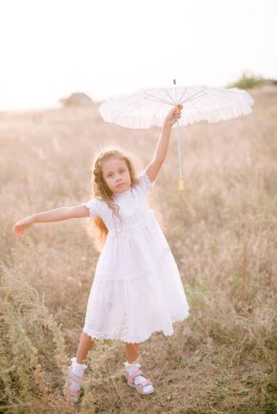 Yazın günbatımında kırsalda beyaz dantel şemsiyeli, uzun sarı kıvırcık saçlı, beyaz bir şapkası olan küçük şirin bir kız. Doğa ve Çevre Yaşamı