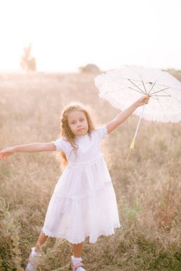 Yazın günbatımında kırsalda beyaz dantel şemsiyeli, uzun sarı kıvırcık saçlı, beyaz bir şapkası olan küçük şirin bir kız. Doğa ve Çevre Yaşamı
