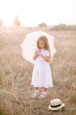 Yazın günbatımında kırsalda beyaz dantel şemsiyeli, uzun sarı kıvırcık saçlı, beyaz bir şapkası olan küçük şirin bir kız. Doğa ve Çevre Yaşamı