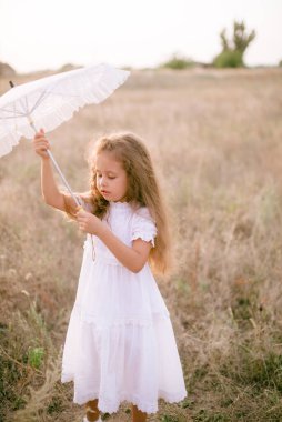 Yazın günbatımında kırsalda beyaz dantel şemsiyeli, uzun sarı kıvırcık saçlı, beyaz bir şapkası olan küçük şirin bir kız. Doğa ve Çevre Yaşamı