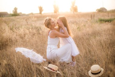 Uzun sarı kıvırcık saçlı şirin bir kız ve yaz günü güneş batarken kırsalda saman kayıkçı şapkalı beyaz elbiseli annesi. Doğa ve Çevre Yaşamı