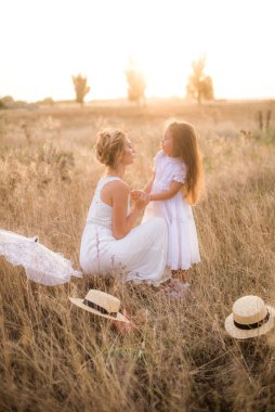 Uzun sarı kıvırcık saçlı şirin bir kız ve yaz günü güneş batarken kırsalda saman kayıkçı şapkalı beyaz elbiseli annesi. Doğa ve Çevre Yaşamı