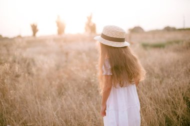 A cute little girl with long blond curly hair and her mother in a white summer dress and a straw boater hat in a field in the countryside in summer at sunset. Nature and Ecolife
