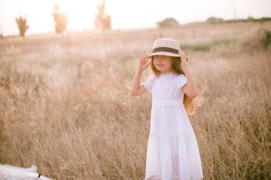 A cute little girl with long blond curly hair and her mother in a white summer dress and a straw boater hat in a field in the countryside in summer at sunset. Nature and Ecolife