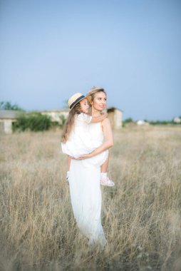 Uzun sarı kıvırcık saçlı şirin bir kız ve yaz günü güneş batarken kırsalda saman kayıkçı şapkalı beyaz elbiseli annesi. Doğa ve Çevre Yaşamı