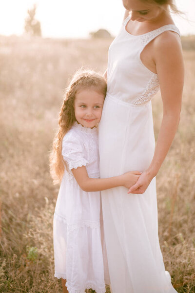 A cute little girl with long blond curly hair and her mother in a white summer dress and a straw boater hat in a field in the countryside in summer at sunset. Nature and Ecolife