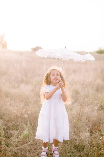 A cute little girl with long blond curly hair in a white summer dress and a straw boater hat with a white lace umbrella in a field in the countryside in summer at sunset. Nature and Ecolife