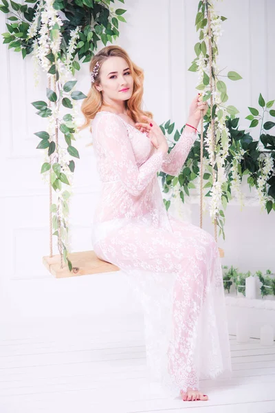 Young attractive bride with blond hair with wedding makeup and hairstyle in a white lace peignoir in a bright interior