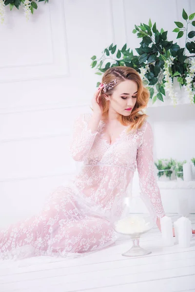 Young attractive bride with blond hair with wedding makeup and hairstyle in a white lace peignoir in a bright interior