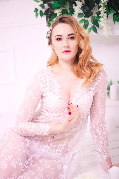 Young attractive bride with blond hair with wedding makeup and hairstyle in a white lace peignoir in a bright interior