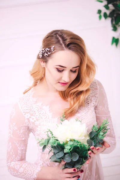 Young attractive bride with blond hair with wedding makeup and hairstyle in a white lace peignoir with bride\'s bouquet in a bright interior