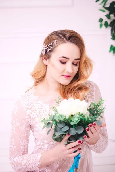 Young attractive bride with blond hair with wedding makeup and hairstyle in a white lace peignoir with bride\'s bouquet in a bright interior