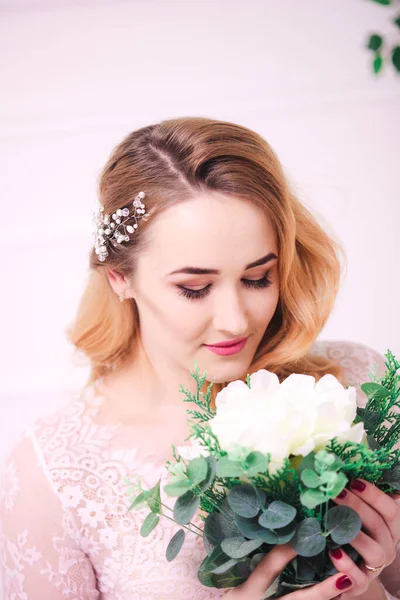 Young attractive bride with blond hair with wedding makeup and hairstyle in a white lace peignoir with bride\'s bouquet in a bright interior