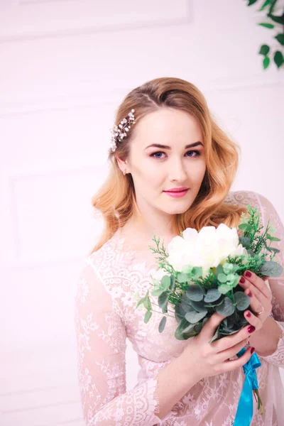 Young attractive bride with blond hair with wedding makeup and hairstyle in a white lace peignoir with bride's bouquet in a bright interior