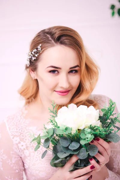 Young attractive bride with blond hair with wedding makeup and hairstyle in a white lace peignoir with bride\'s bouquet in a bright interior