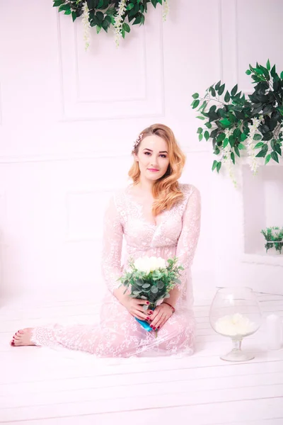 Young attractive bride with blond hair with wedding makeup and hairstyle in a white lace peignoir with bride\'s bouquet in a bright interior