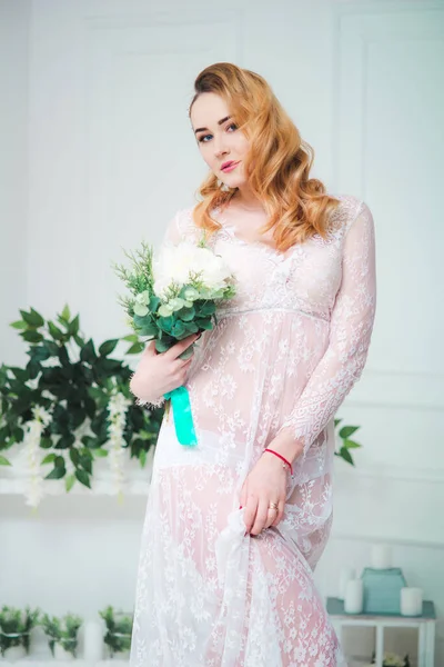 Young attractive bride with blond hair with wedding makeup and hairstyle in a white lace peignoir with bride\'s bouquet in a bright interior