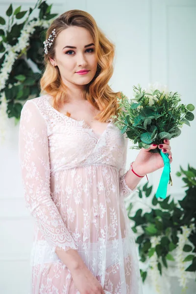 Young attractive bride with blond hair with wedding makeup and hairstyle in a white lace peignoir with bride\'s bouquet in a bright interior