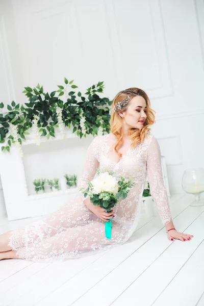 Young attractive bride with blond hair with wedding makeup and hairstyle in a white lace peignoir with bride\'s bouquet in a bright interior