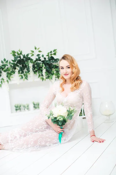 Young attractive bride with blond hair with wedding makeup and hairstyle in a white lace peignoir with bride's bouquet in a bright interior