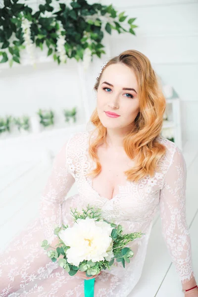 Young attractive bride with blond hair with wedding makeup and hairstyle in a white lace peignoir with bride\'s bouquet in a bright interior