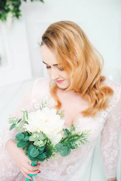 Young attractive bride with blond hair with wedding makeup and hairstyle in a white lace peignoir with bride\'s bouquet in a bright interior