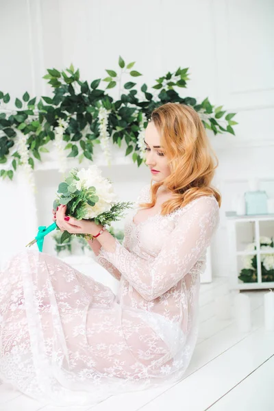 Young attractive bride with blond hair with wedding makeup and hairstyle in a white lace peignoir with bride's bouquet in a bright interior