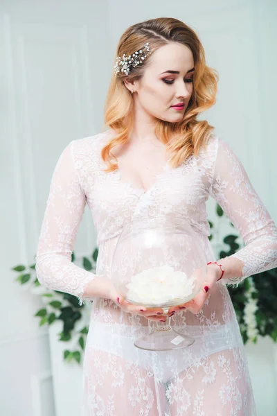 Young attractive bride with blond hair with wedding makeup and hairstyle in a white lace peignoir in a bright interior